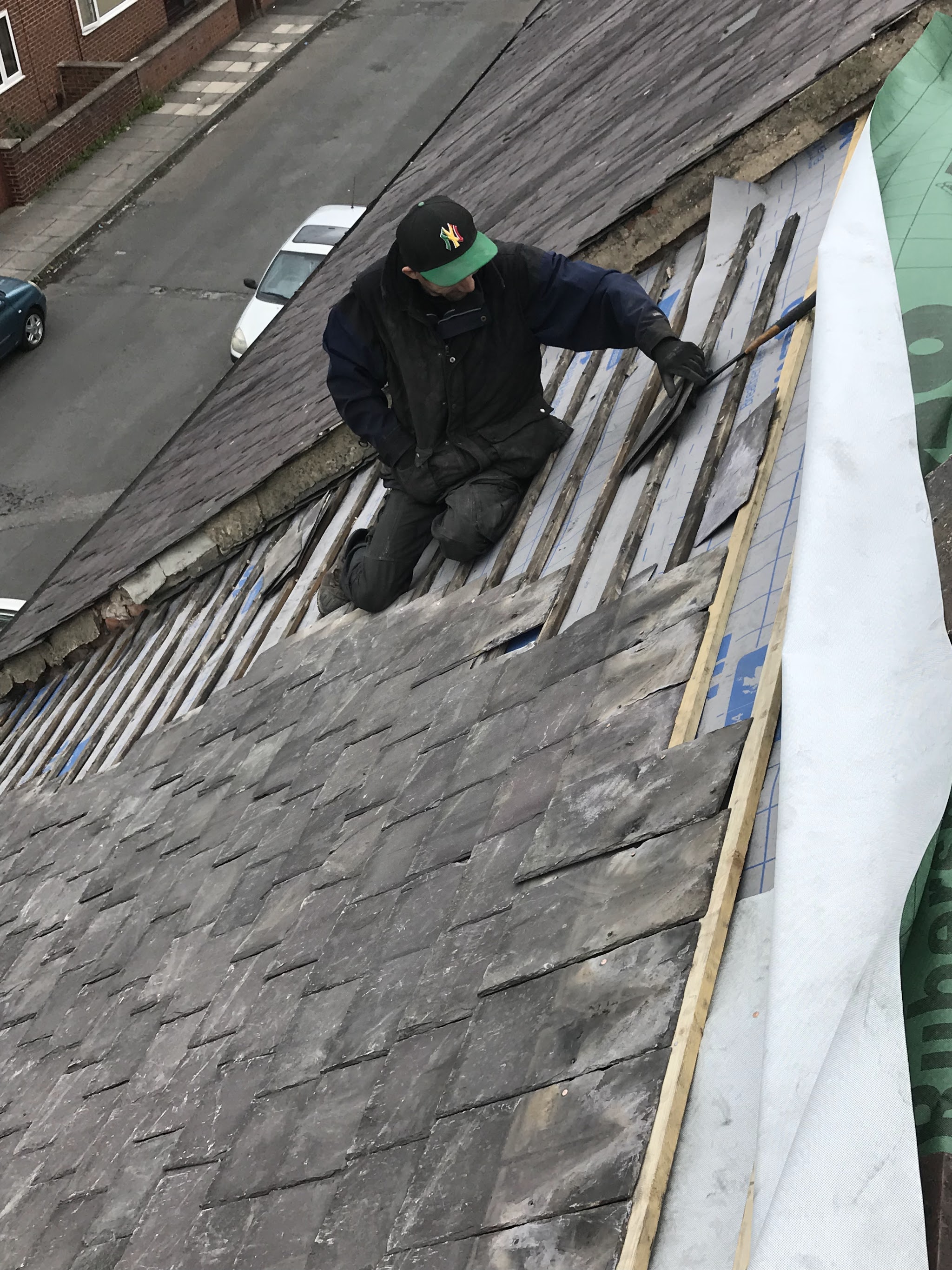 A roofer in a cap kneeling on the roof, laying slates over new battens and membrane above a terraced street